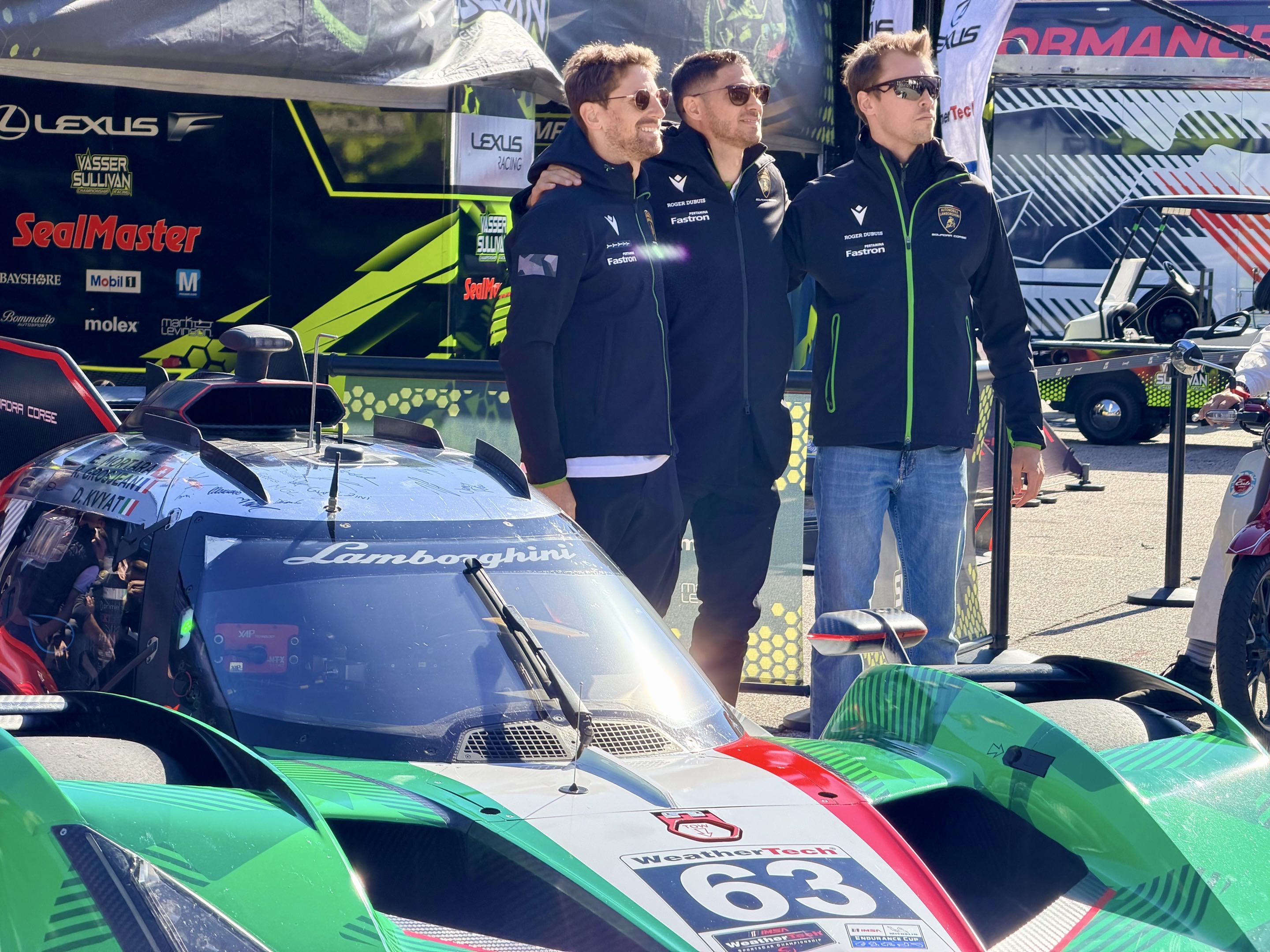 Three racing drivers in jackets pose for a photo next to a Lamborghini SC63 prototype race car
